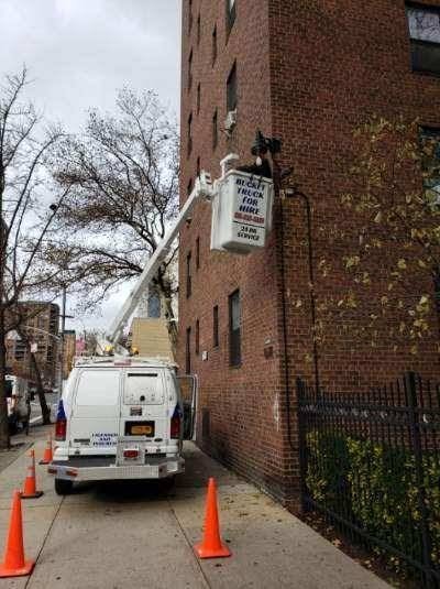 Our van on the sidewalk with the bucket raised beside the upper floors of a brick apartment building, safety cones around the truck